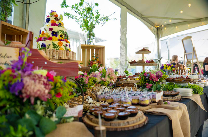table filled with various food platters and natural burlap and flower decor, highlighted by a white cake adorned with colorful wildflowers