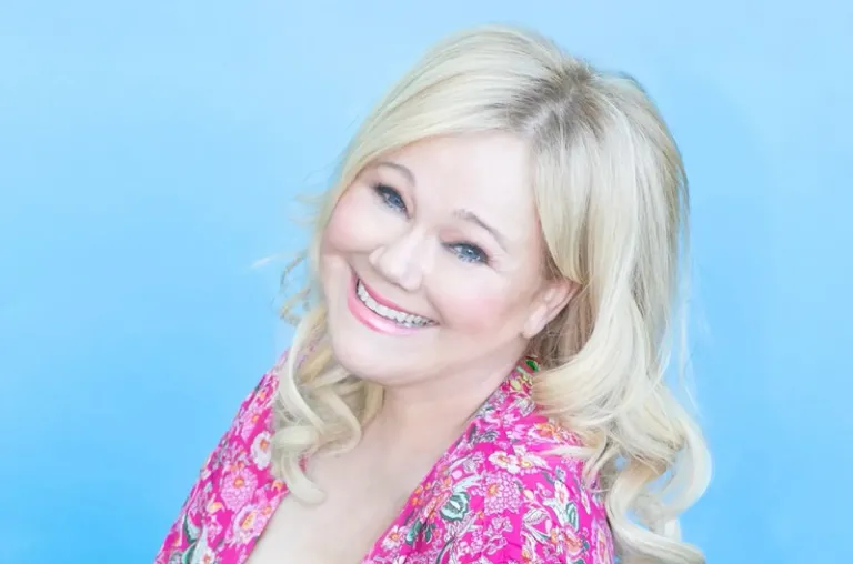 A blonde woman in a pink print shirt agains a light blue background